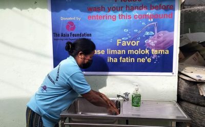 woman in face mask washes her hand in front of banner that encourages handwashing